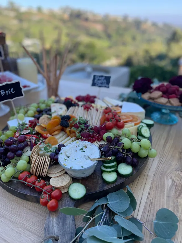A round wooden board of grapes, crackers, whipped dip, cucumber and cherry tomatoes trimmed with eucalyptus