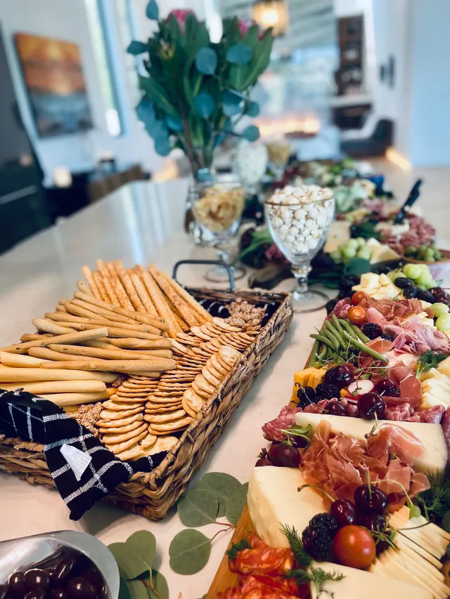 An indoor grazing spread with a basket of breadsticks and crackers, popcorn in a glass and prosciutto with cherries