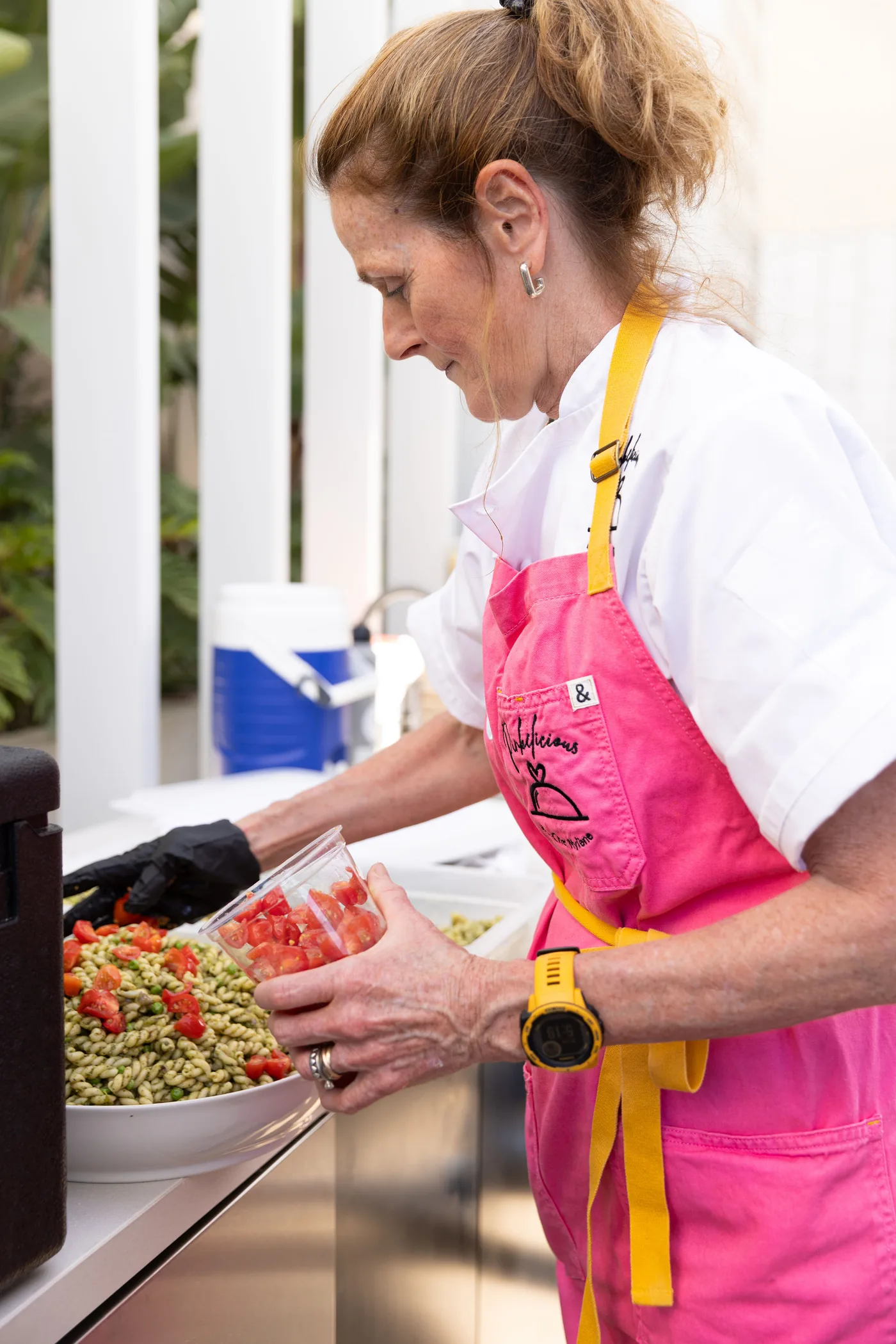 Chef Mylène in her pink apron adding chopped tomatoes to a bowl of pesto pasta salad