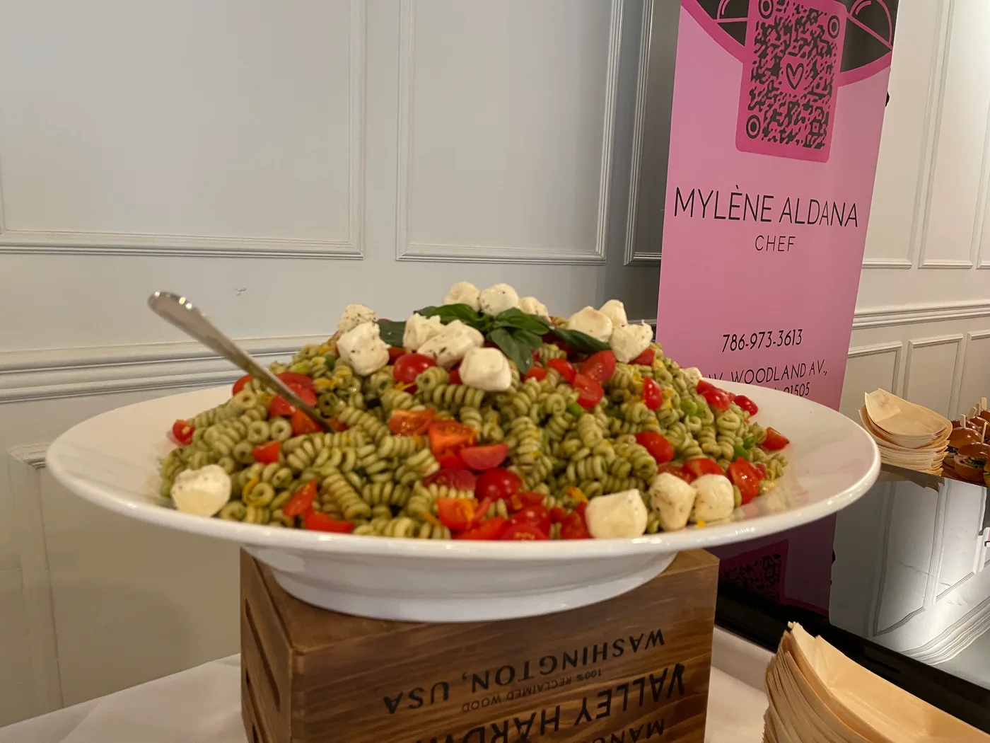 A bowl of pesto pasta salad with tomatoes and mozzarella displayed on a wooden crate